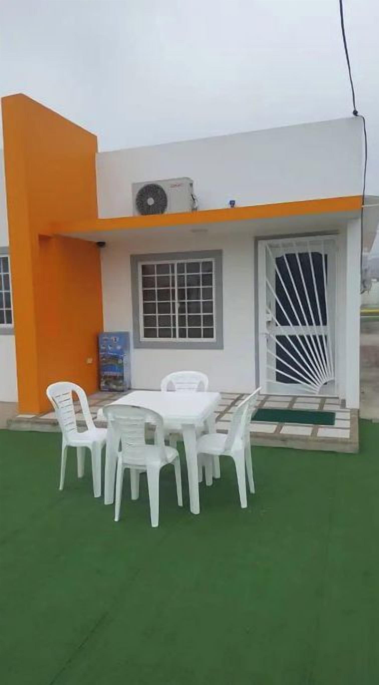 Un bâtiment à la façade blanche et orange, doté d’un espace extérieur abrité avec des chaises en plastique blanc et une table, ainsi que d’une pelouse artificielle verte.