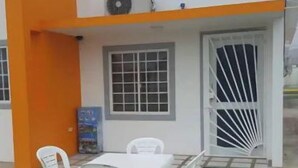 Ein Gebäude mit weiß-orangener Fassade, ein überdachter Außenbereich mit weißen Plastikstühlen und einem Tisch sowie eine grüne Rasenfläche.