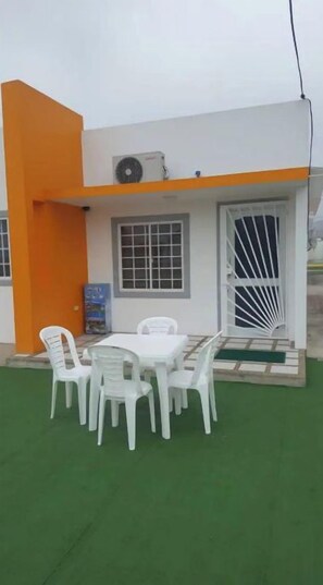 A building with a white and orange exterior, a covered outdoor seating area with white plastic chairs and a table, and a green artificial lawn.