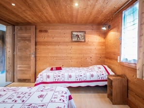 Ein Zimmer in einer Holzhütte mit zwei Betten, einer Holzbalkendecke, einem gerahmten Bild an der Wand und einem Fenster mit Vorhängen.