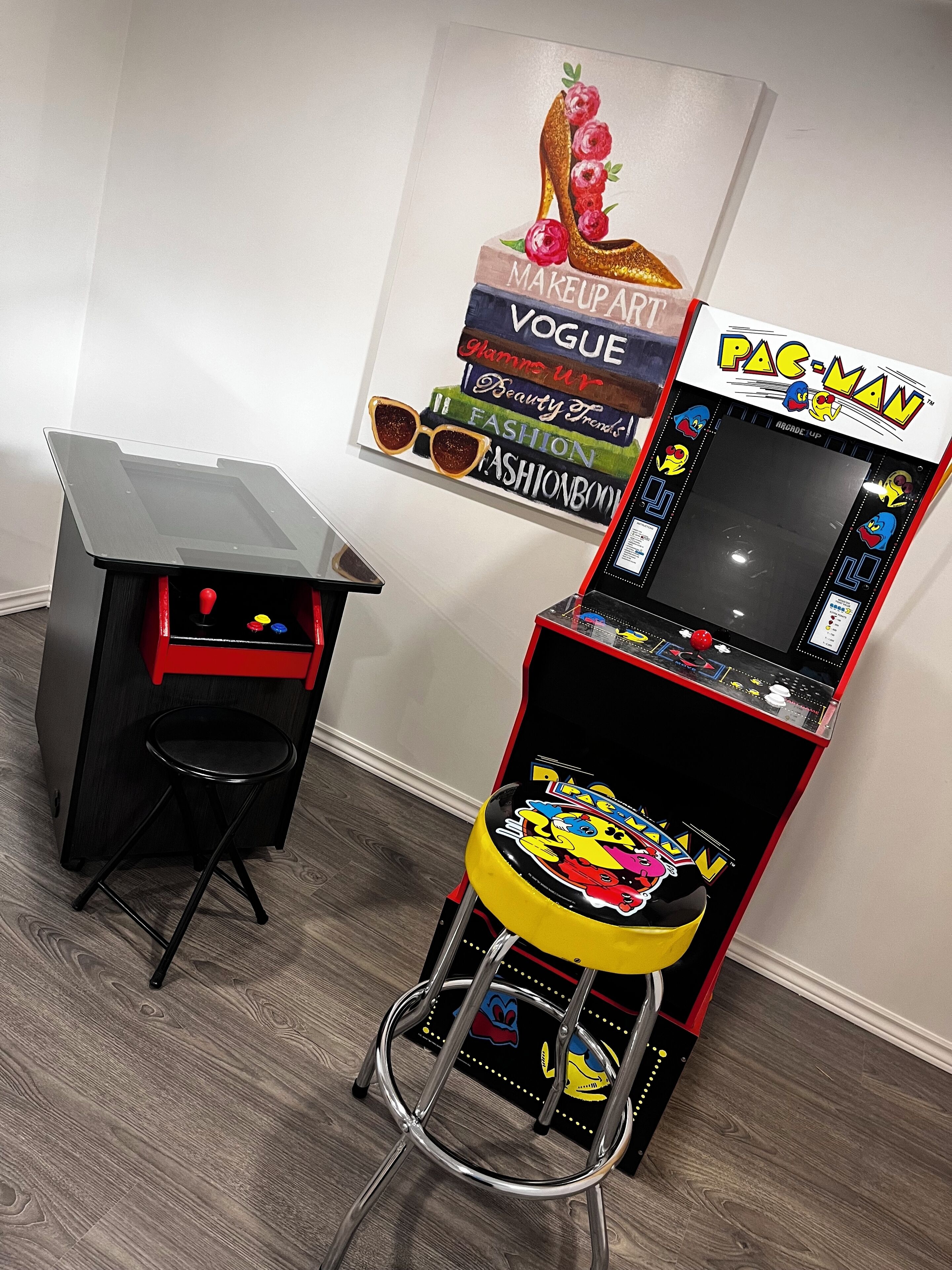 Une salle d’arcade avec des jeux comme Pac-Man et d’autres jeux classiques, un petit tabouret et une affiche accrochée au mur.