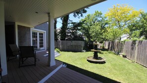 Eine überdachte Terrasse mit Holzdeck, einer Sitzgruppe mit Stühlen, einem kleinen Brunnen und einem gepflegten Rasen.