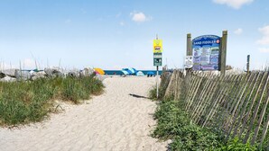 Ein sandiger Pfad führt zu einem Strandbereich mit einem Schild und badenden Personen in der Ferne.