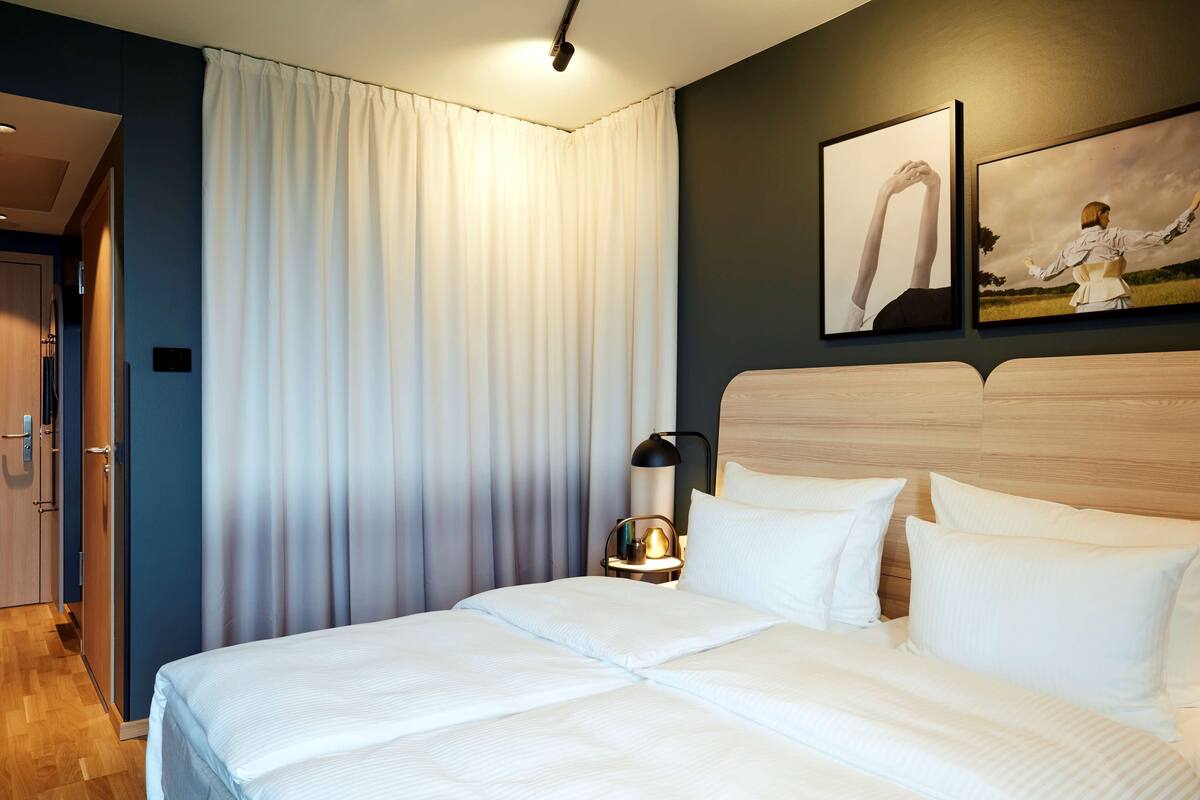 A neatly made bed with white linens, a bedside table with a lamp, and a large window with curtains.