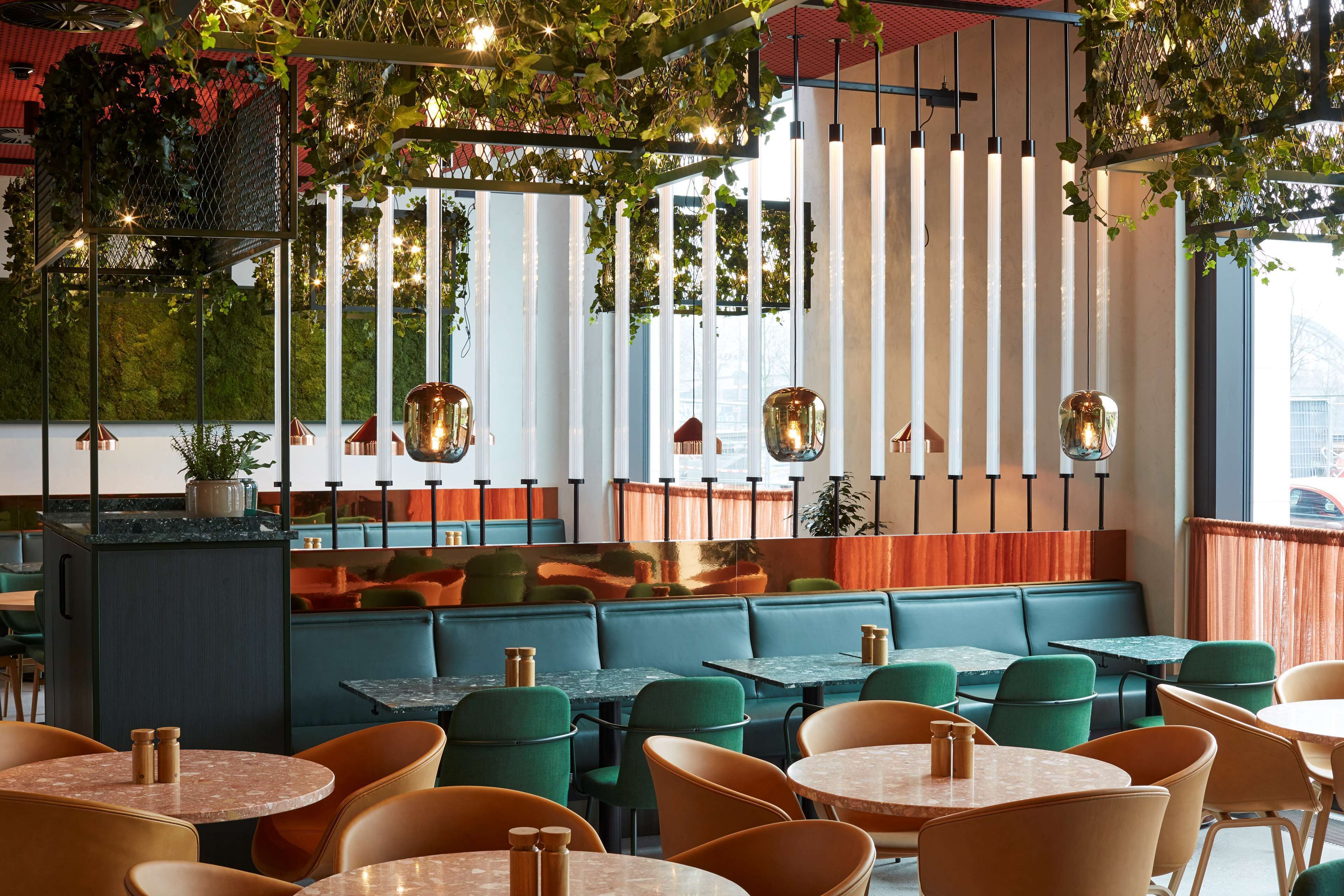 Een restaurant met zitjes en tafels, groene en oranje stoelen, hangplanten en hanglampen.