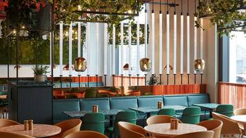 Een restaurant met zitjes en tafels, groene en oranje stoelen, hangplanten en hanglampen.