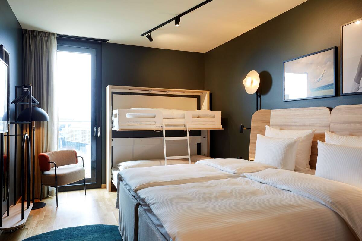 A hotel room with a bunk bed, a desk with a chair, a standing lamp, and a large window.