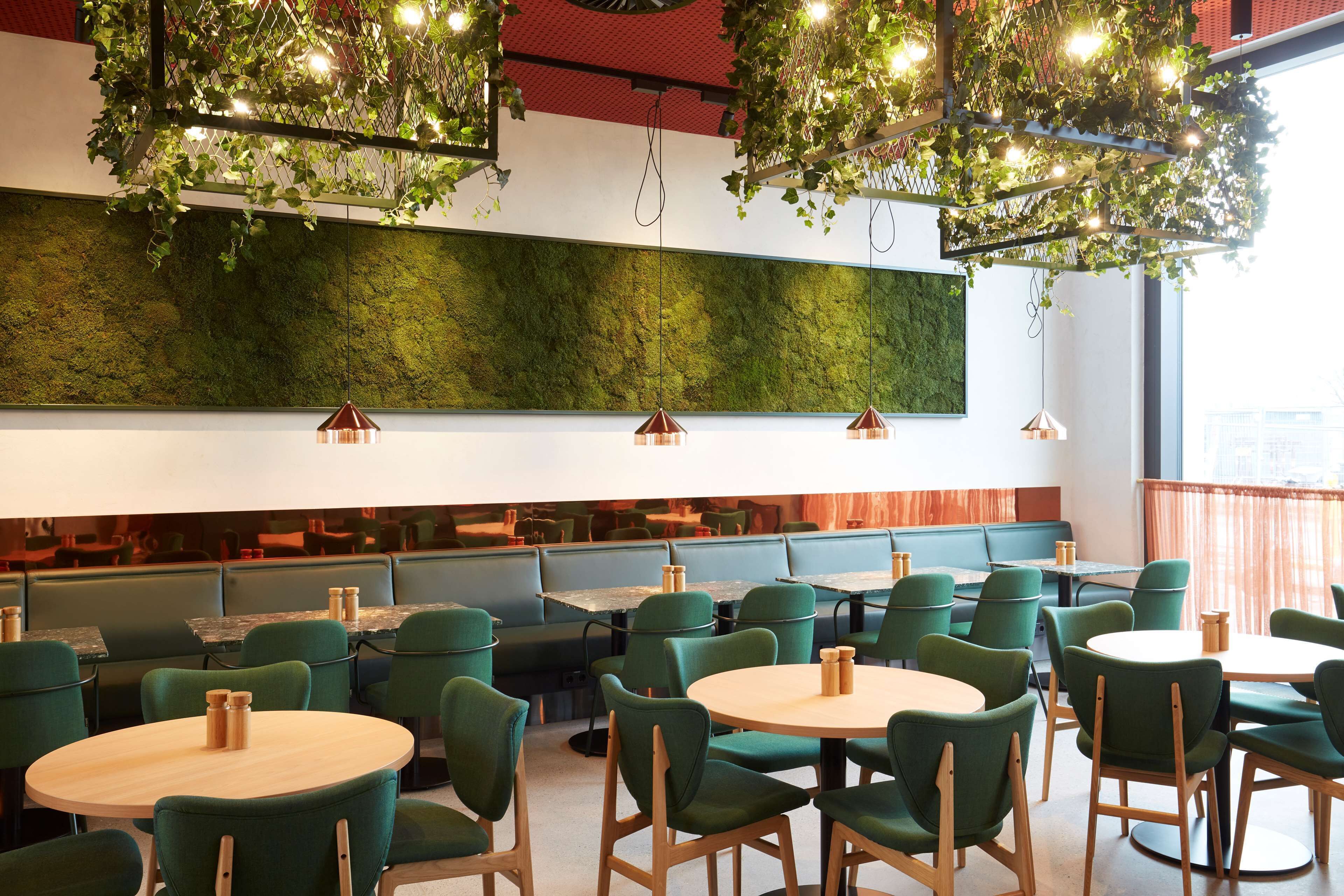 A restaurant with green upholstered booths, round tables, and hanging light fixtures with greenery.