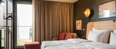 Habitación de hotel con una cama grande, una zona de estar con un cojín rojo, vistas de la ciudad a través de un ventanal amplio y una lámpara fijada a la pared.