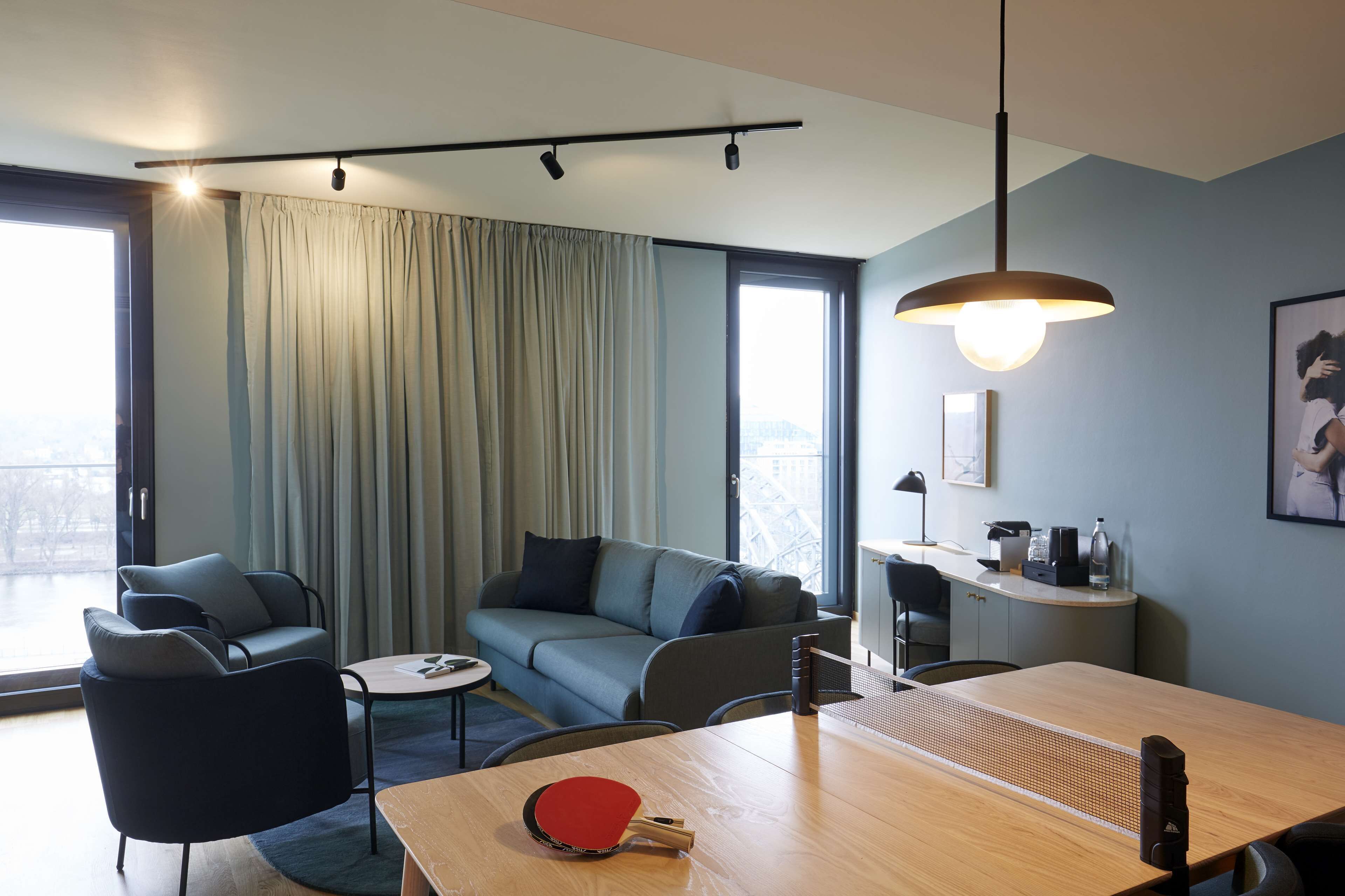 Habitación de hotel moderna con zona de comedor, sofá, mesa con un tazón rojo y vistas a la ciudad a través de ventanas amplias.
