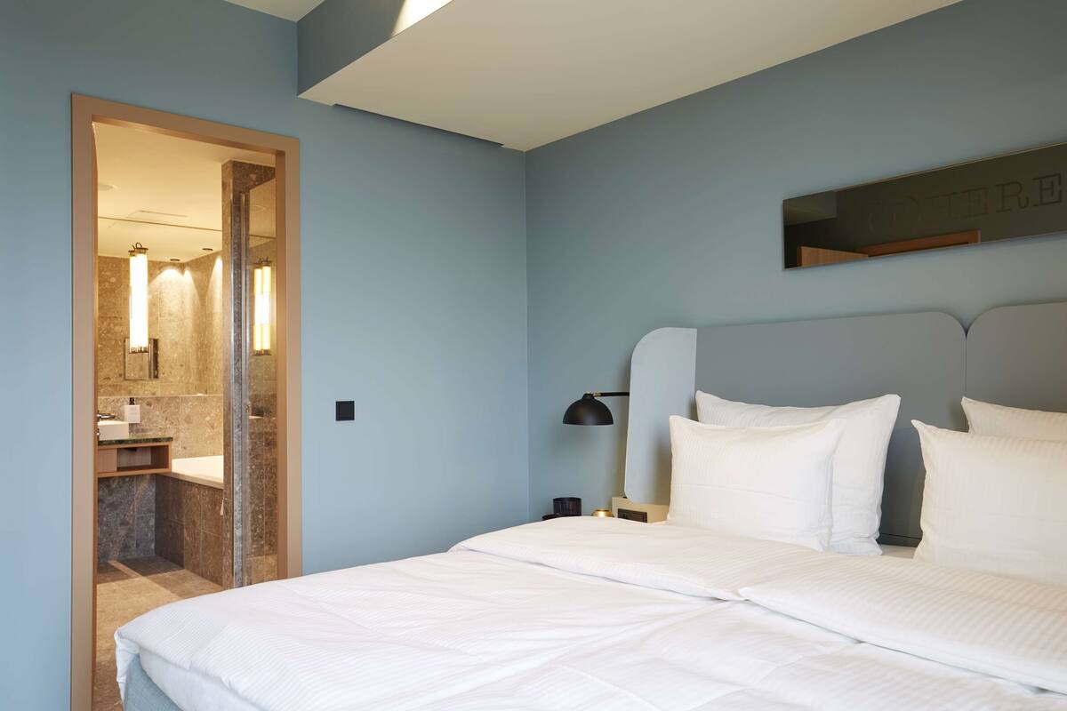 A bedroom with a large bed, a nightstand with a lamp, a wall-mounted mirror, and a bathroom visible through an open door.