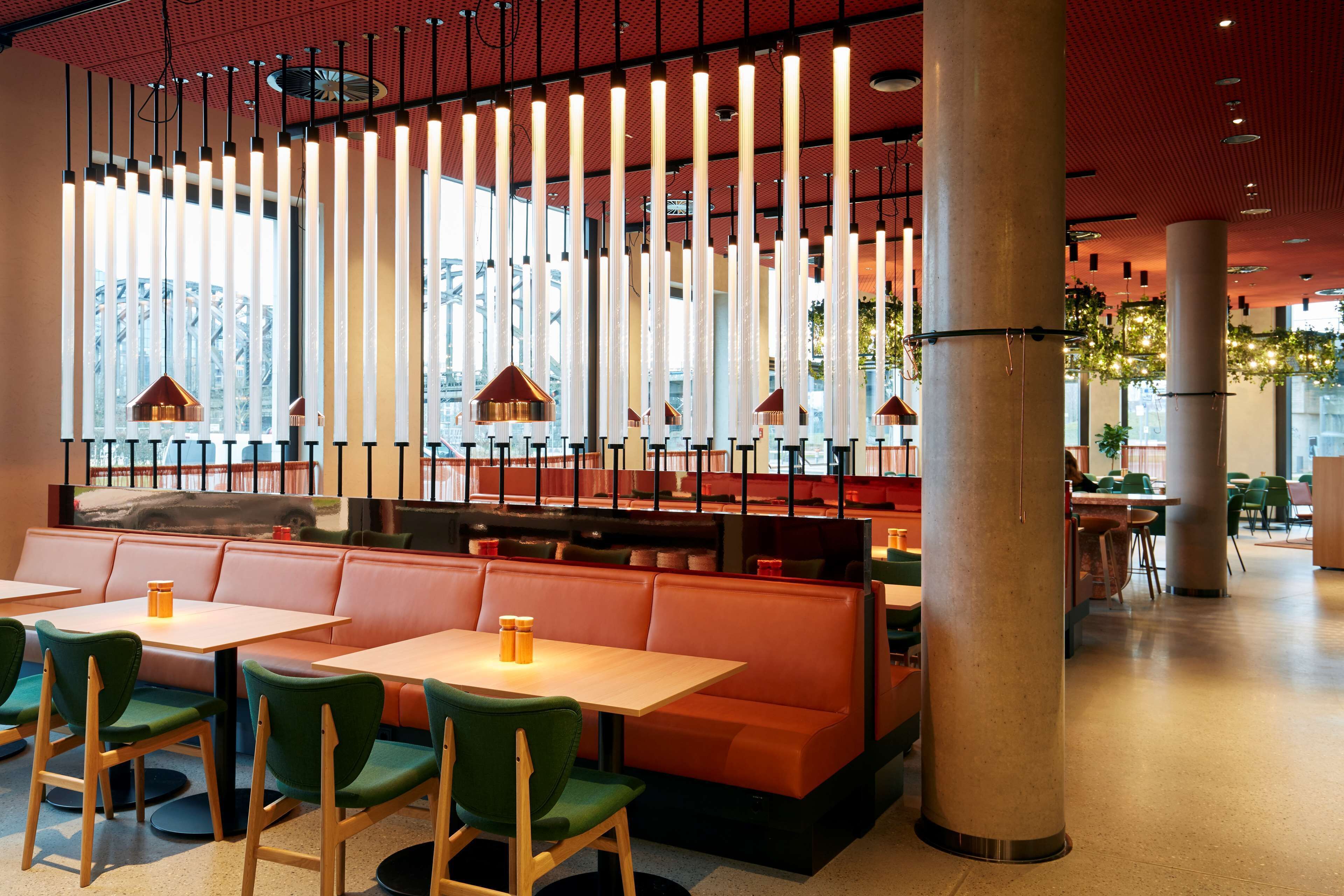 Interior moderno de restaurante con techos altos, lámparas verticales y una variedad de opciones de asientos, incluyendo cabinas y mesas.