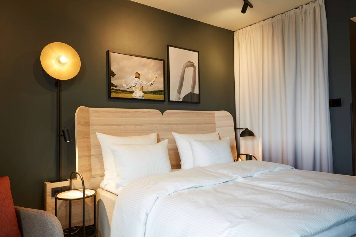 A hotel room with a bed, bedside lamps, a nightstand, a chair, and artwork on the wall.