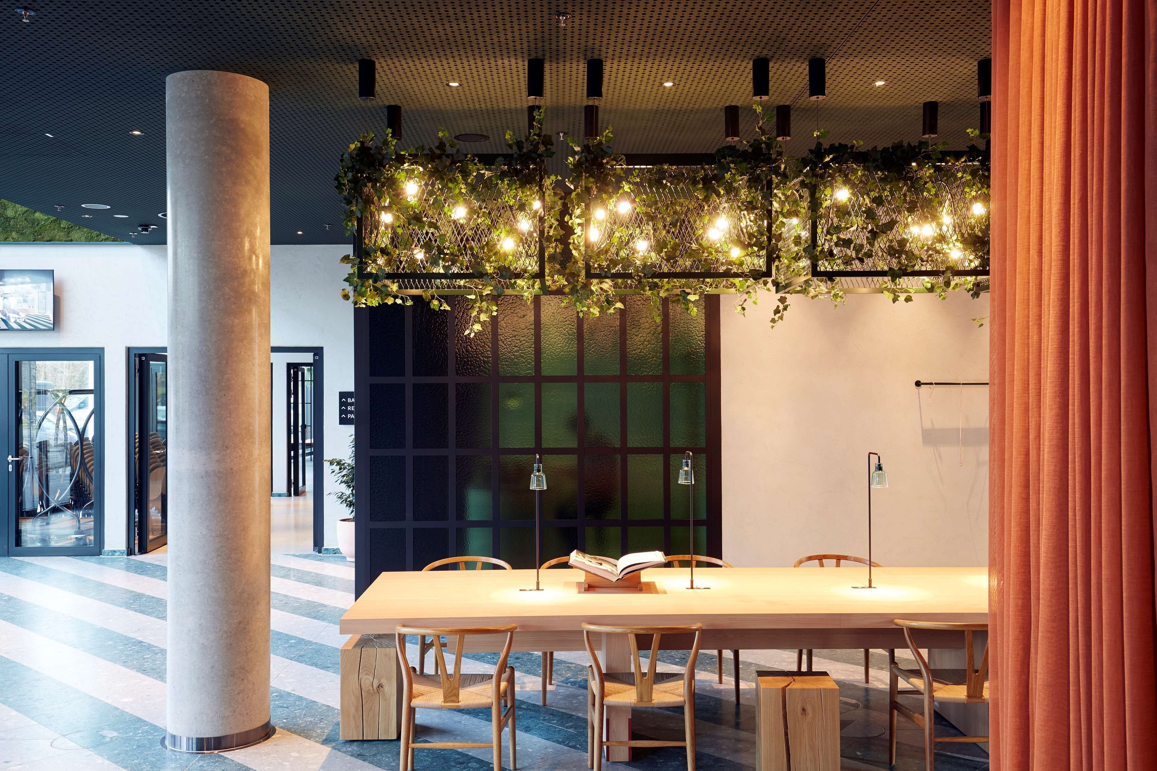 Un espacio interior moderno con una mesa grande, sillas, una columna y una lámpara colgante con plantas.