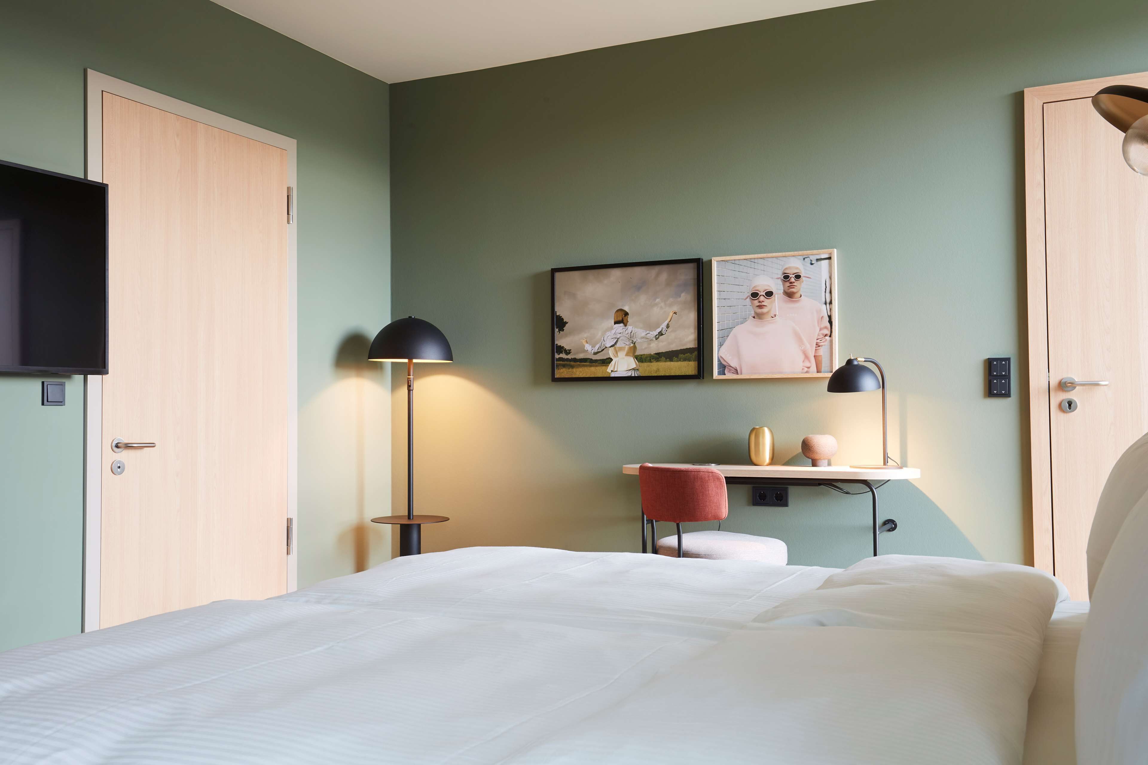 A bedroom with a bed, a desk with a chair, a lamp, and two framed pictures on the wall.