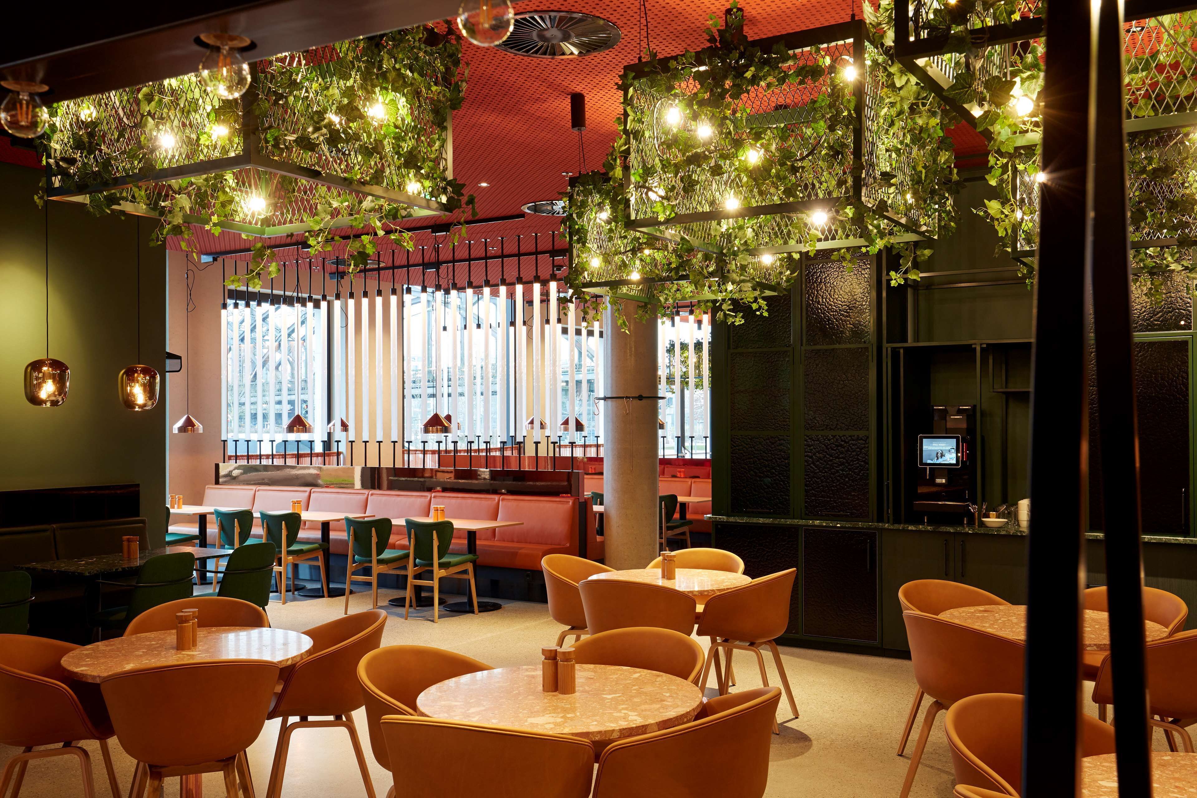 A restaurant with round tables and orange chairs, a bar area with a TV, and green plants hanging from the ceiling.
