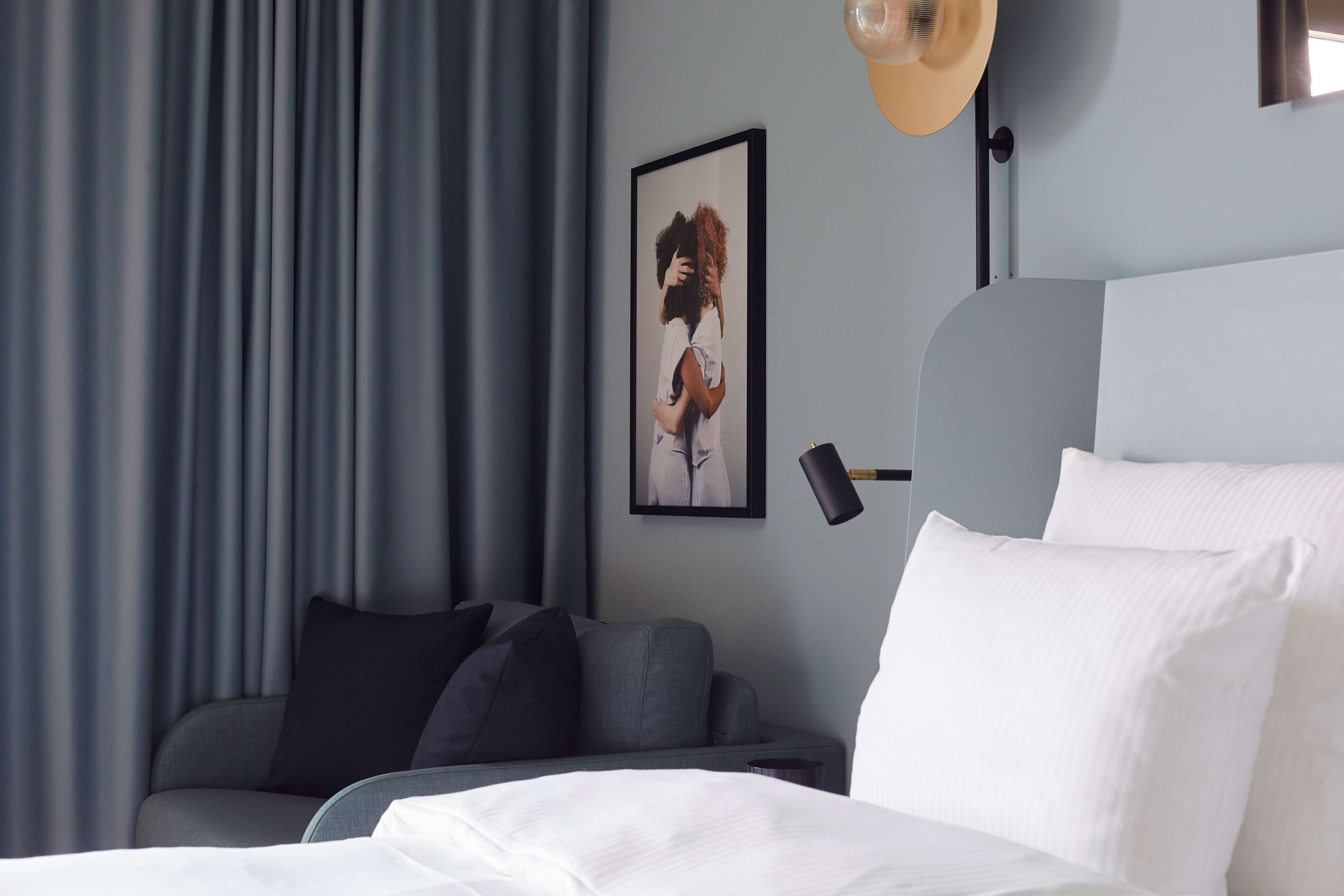 A modern hotel room with a bed, a sofa, a wall-mounted lamp, and a framed picture.