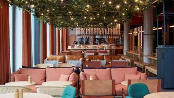 A restaurant with multiple seating areas, including round tables and cushioned booths, under a canopy of greenery and string lights.