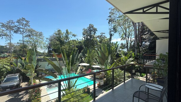 Un balcón con vista a una piscina, rodeado de plantas tropicales y un coche estacionado.