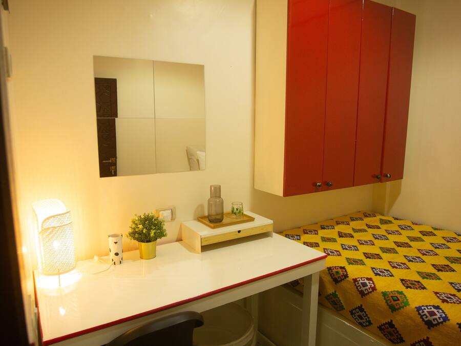 A small, well-lit bedroom with a desk, a bed with a patterned bedspread, a red wardrobe, and a mirror on the wall.