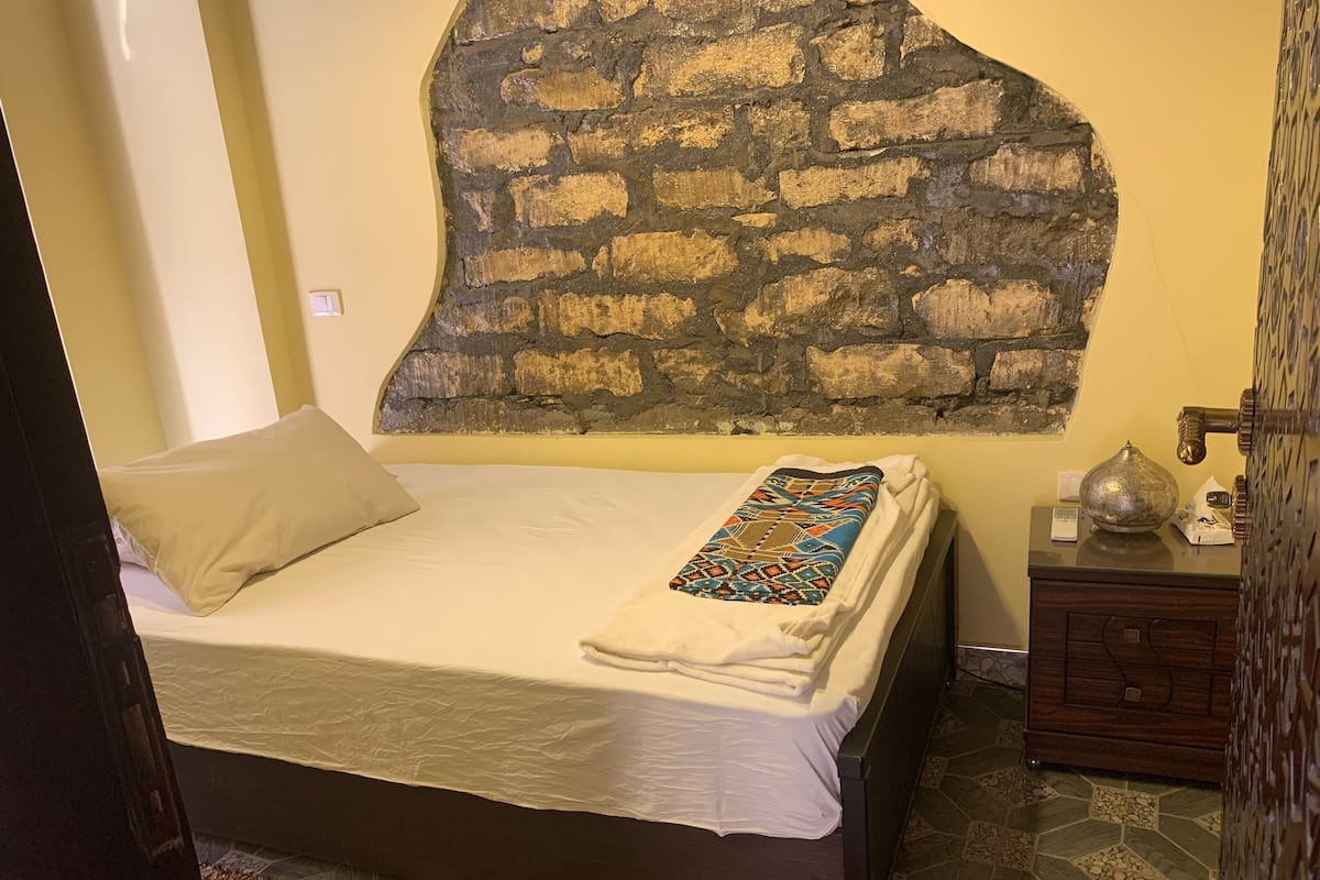 Cama bem arrumada com manta estampada, mesa de cabeceira com objeto decorativo e parede de pedra.