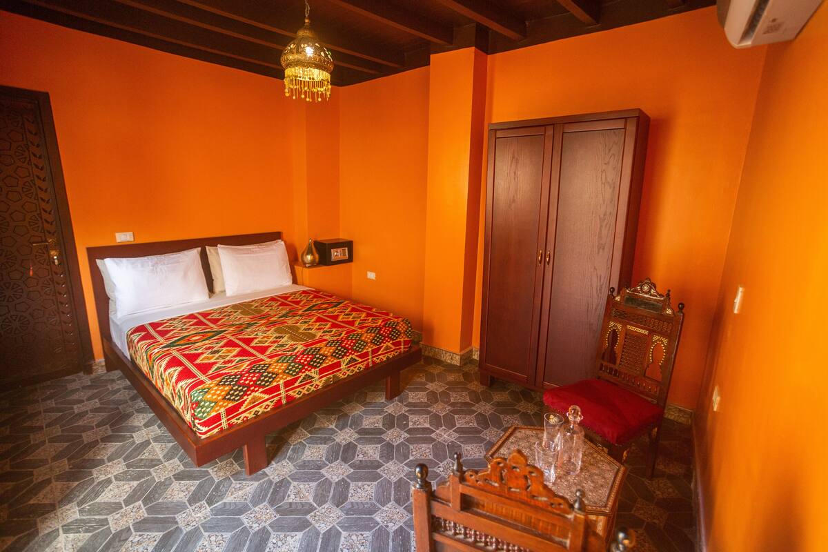 Quarto com cama grande, guarda-roupa de madeira, candelabro e pequena mesa com almofada vermelha.