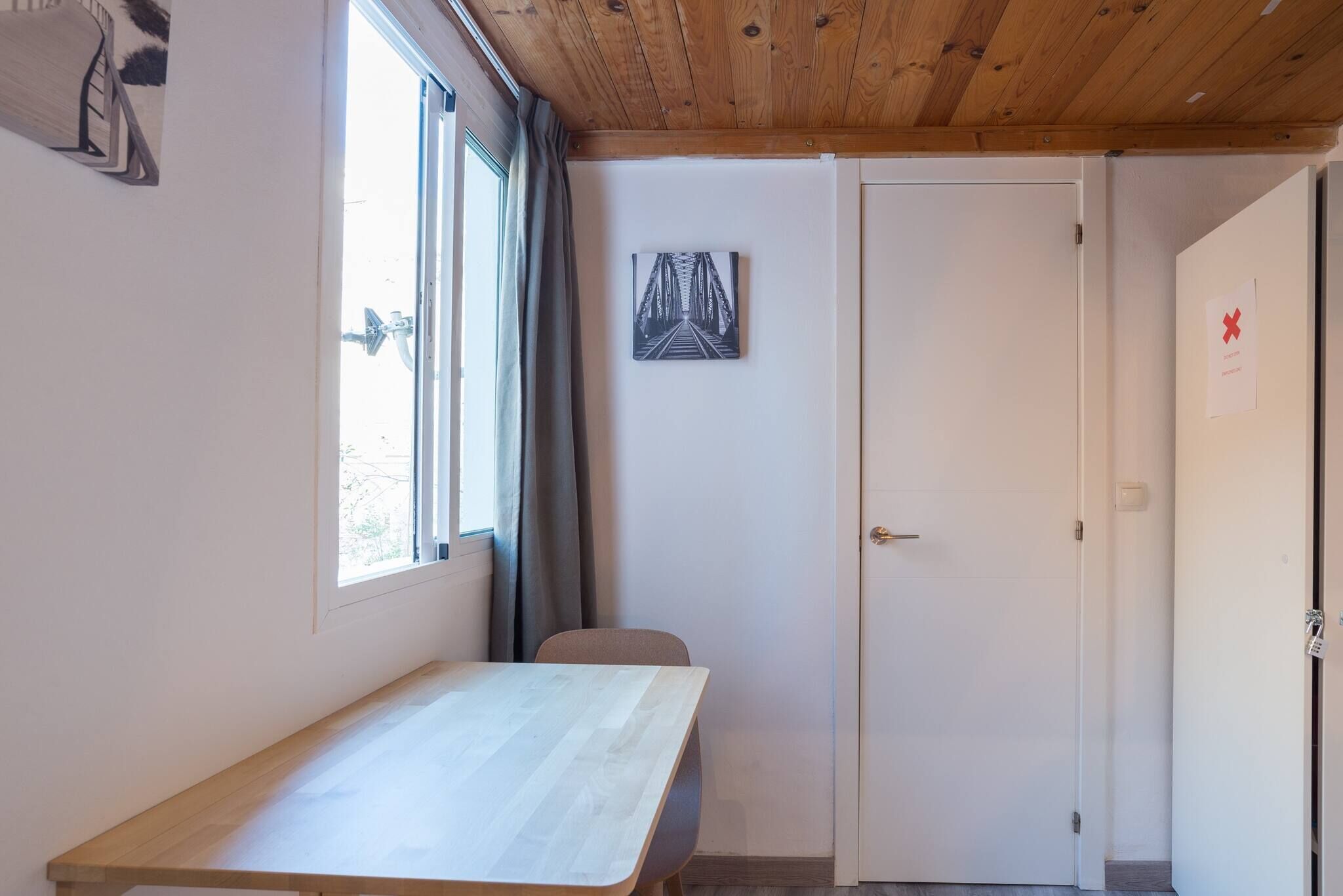 Una habitació amb sostre de fusta, una finestra amb cortines, una porta blanca i un quadre encimbellat a la paret.