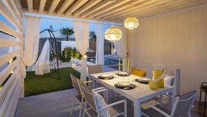 A dining area with a white table set for four, outdoor seating, and a pool in the background.