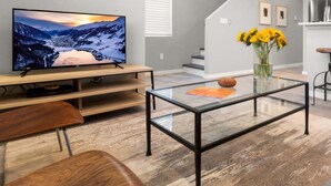 Un salon moderne avec un téléviseur à écran plat, une table basse en verre, des meubles en bois et un coin cuisine à l’arrière-plan.