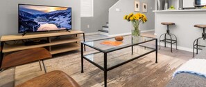A modern living room with a flat-screen TV, a glass coffee table, wooden furniture, and a kitchen area in the background. - Downtown Luxury Townhouse w/Garage & Walkable (Albuquerque)