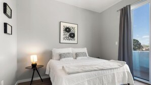 Quarto com uma cama grande, mesa de cabeceira com abajur, quadro na parede e vista para o exterior por uma porta deslizante.