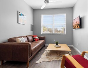 A living room with a brown leather sofa, a wooden coffee table, a red chair, a white rug, and a large window with blinds.