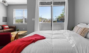 A bedroom with a large bed, a red throw blanket, a brown leather sofa, and a window with blinds.