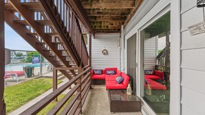 Un espace extérieur aménagé, abrité, avec des canapés rouges et une table en verre, des rampes en bois et une vue sur un bâtiment en arrière-plan.