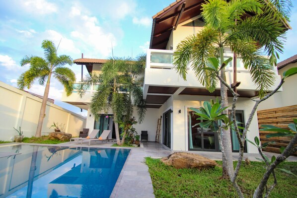 A modern two-story house with a swimming pool, surrounded by tropical plants and a wooden deck.