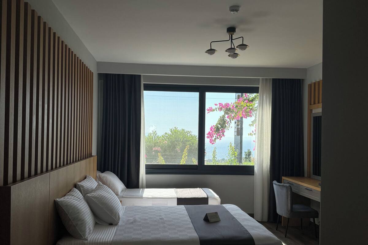 A modern hotel room with a large bed, a desk, and a view of greenery and flowers through the window.