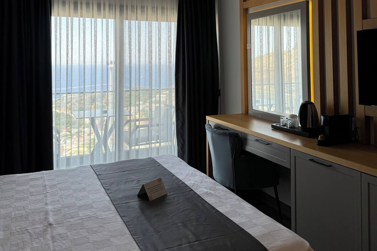 A hotel room with a bed, a desk, a chair, a television, and a view of the sea.
