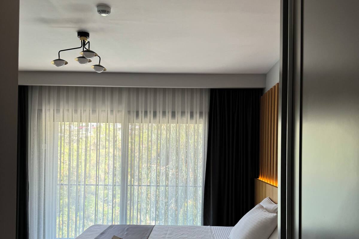 A modern hotel room with a large bed, a ceiling light, and a view through sheer curtains.