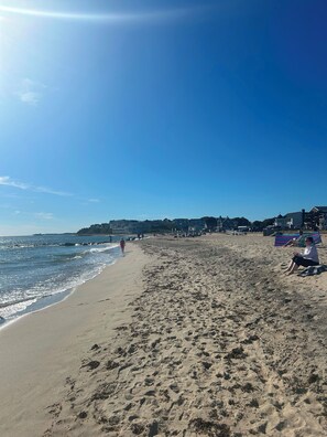 En strand med fodspor, en klar himmel og bygninger i baggrunden.