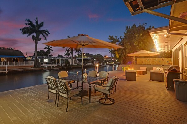 An outdoor deck with a dining table and chairs, surrounded by umbrellas, overlooking a body of water with boats and a dock in the background.