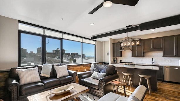 A modern living room with a large window, a ceiling fan, and a kitchen area with dark cabinets and stainless steel appliances.