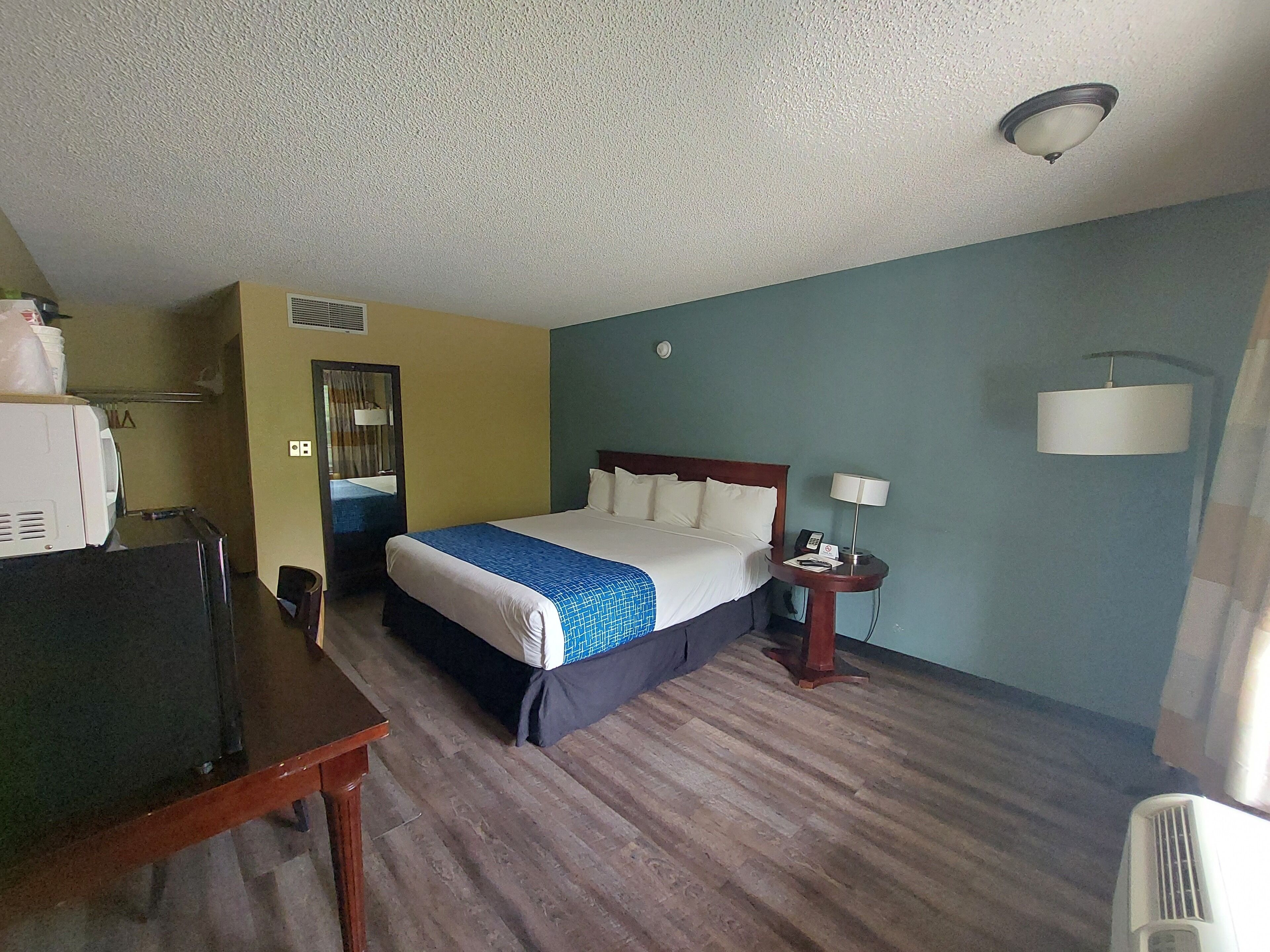 A hotel room with a bed, a desk, a chair, a mirror, a nightstand, and a lamp.