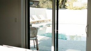 Un salon avec un canapé et une vue, grâce à une porte-fenêtre coulissante, sur un espace extérieur comprenant une piscine et des chaises longues.