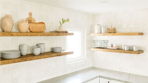 Une cuisine aux murs recouverts de carreaux blancs, avec des étagères en bois où sont rangés les ustensiles de cuisine, et une fenêtre qui laisse entrer la lumière naturelle.