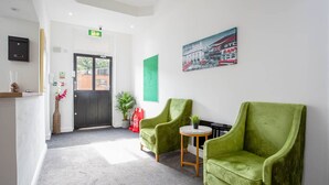 Une salle d’attente avec une porte noire, deux fauteuils verts, une petite table ronde, une plante en pot et un tableau encadré au mur.