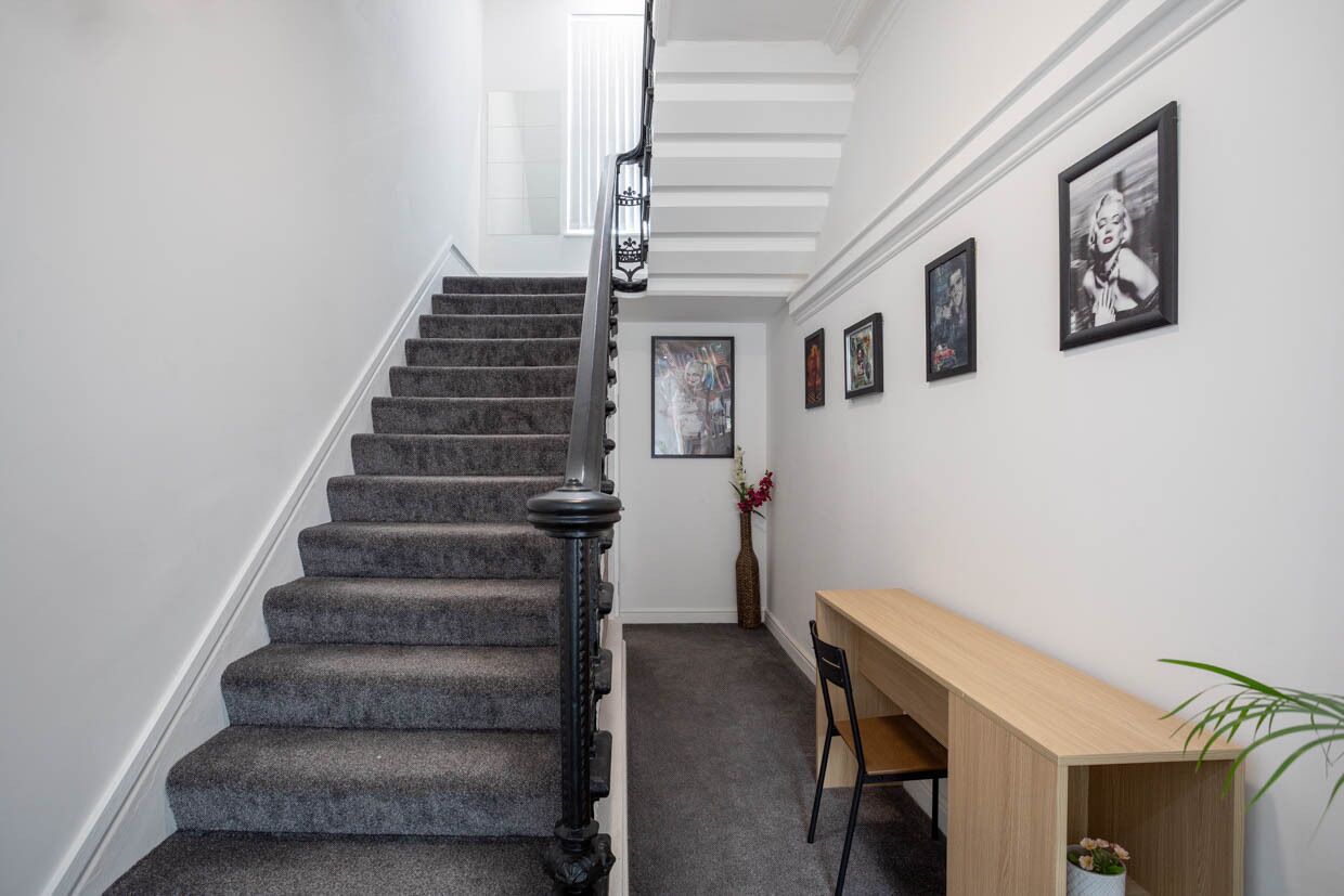 A staircase with a dark carpet and black balusters, leading to a landing with a wooden desk and chair, and framed pictures on the wall.