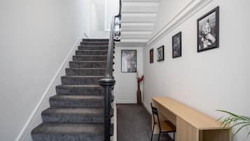 A staircase with a dark carpet and black balusters, leading to a landing with a wooden desk and chair, and framed pictures on the wall.