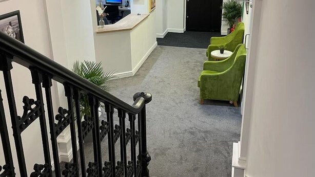 Un hall d’hôtel avec un comptoir d’accueil, un escalier et des fauteuils verts.