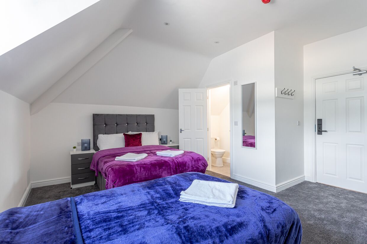 Ein Schlafzimmer mit zwei Betten, einem grauen Kopfstück, einer weißen Tür und einer weißen Wand mit Spiegel.