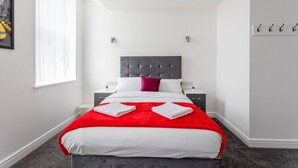 Une chambre d’hôtel moderne avec un grand lit, un plaid rouge et une tête de lit grise. On y trouve deux tables de chevet avec des lampes et une fenêtre avec des stores.